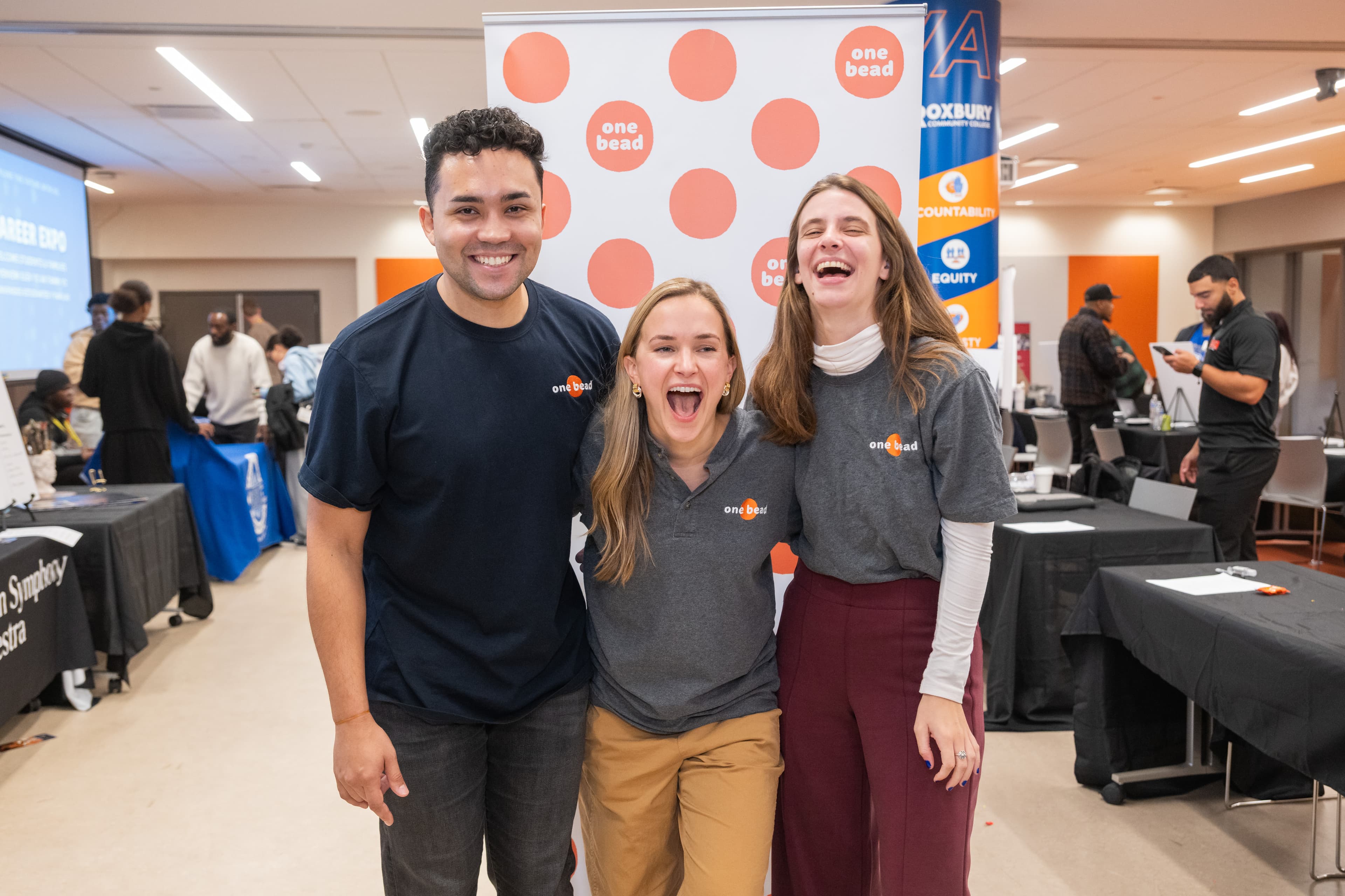 Three people laughing together at a step-and-repeat, in matching branded shirts, career expo tables behind them.