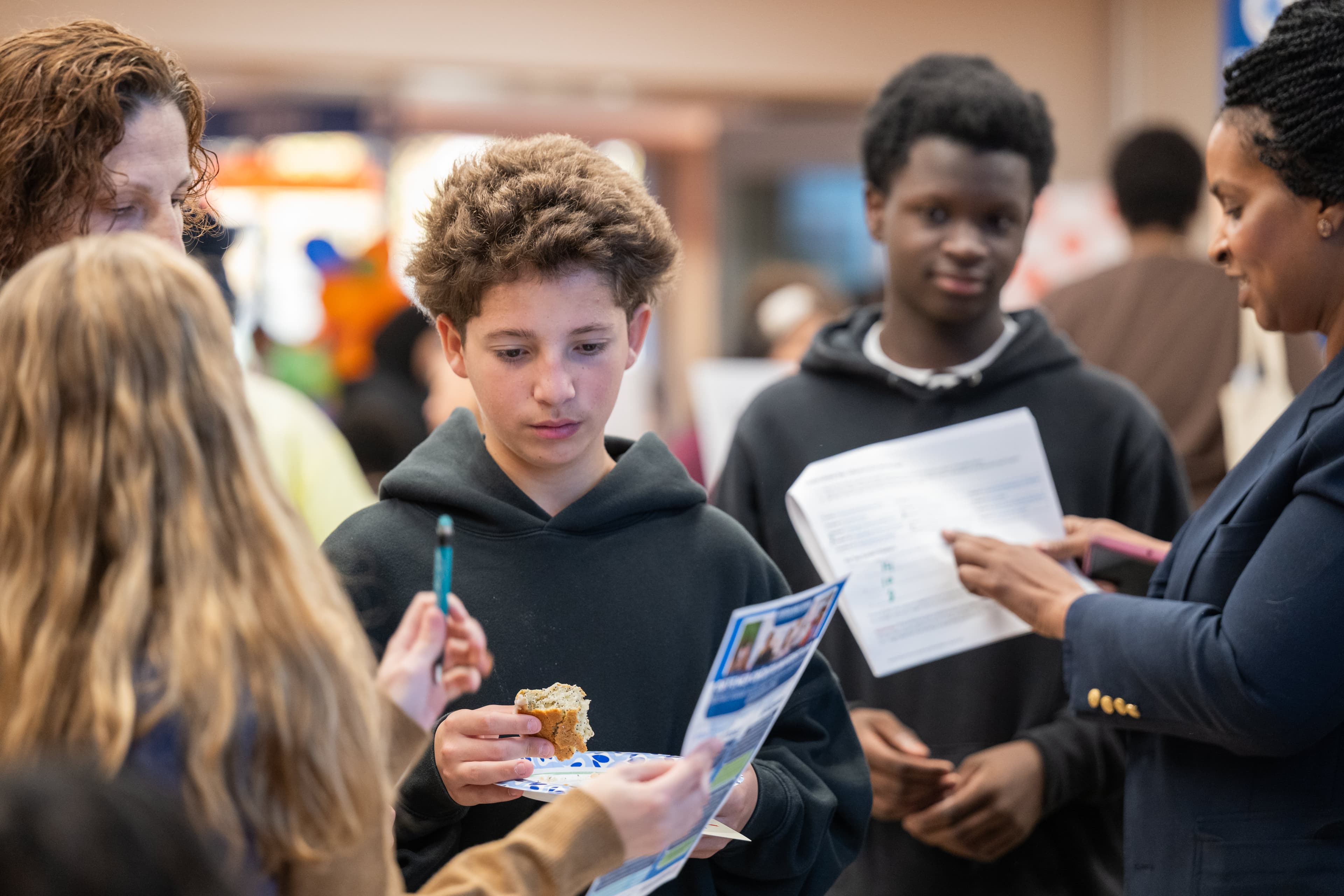 Teenage boy holding a plate with bread, looking at a pamphlet, other teens and an adult reviewing papers nearby.