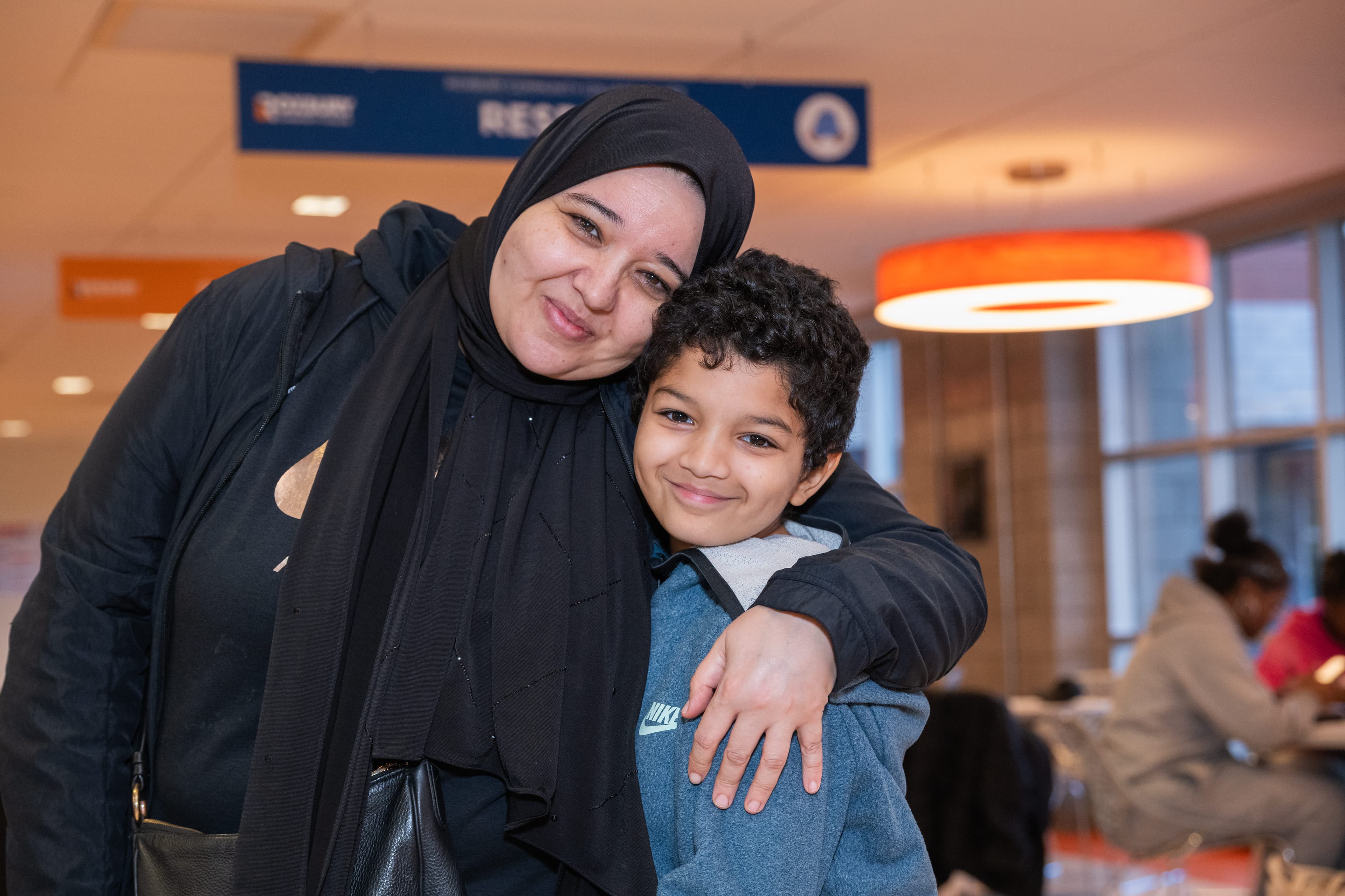 Woman in a hijab embracing a young boy warmly, both smiling at the camera, event signage overhead.