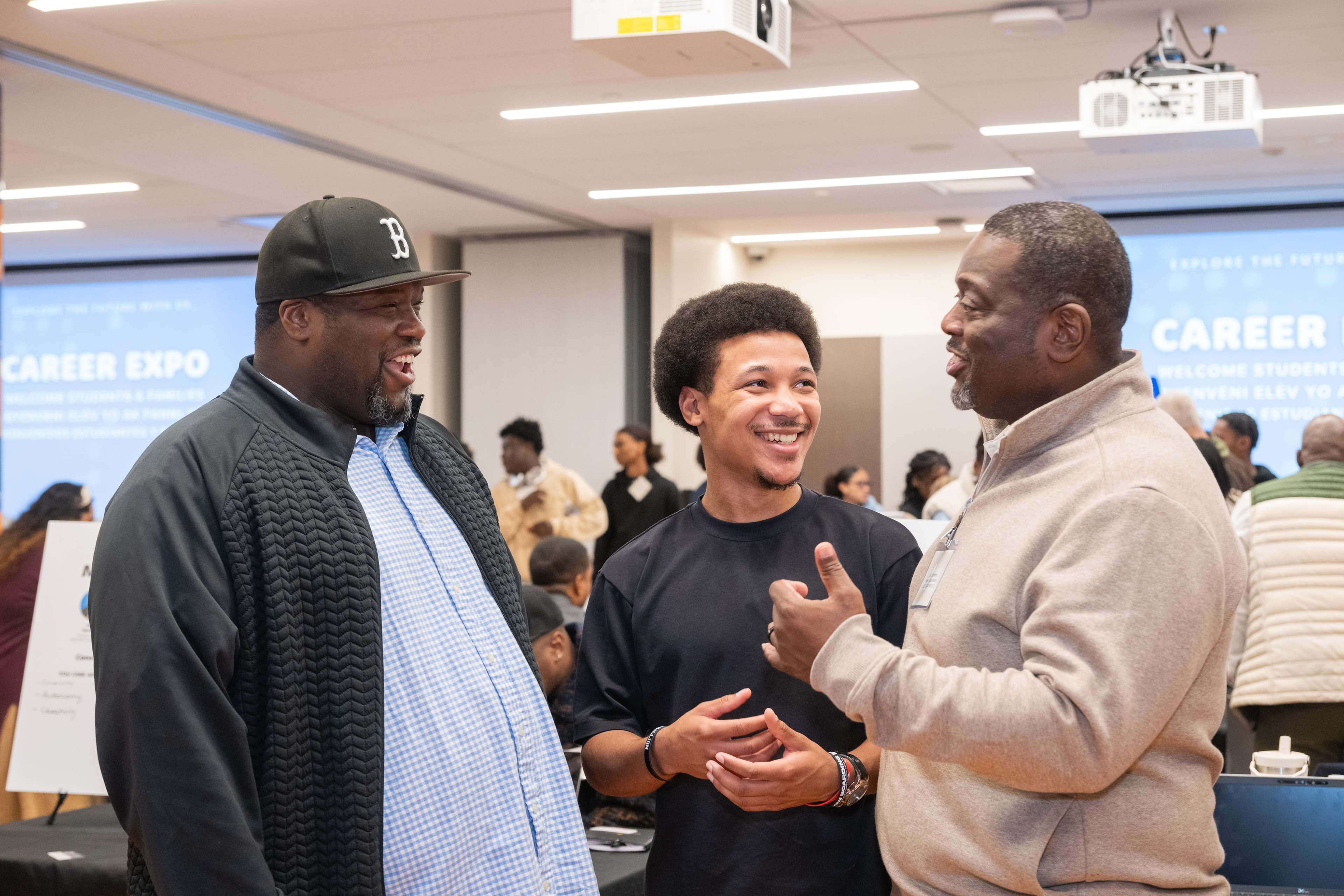 Three men talking and laughing together, one wearing a baseball cap, presentation screens visible behind them.