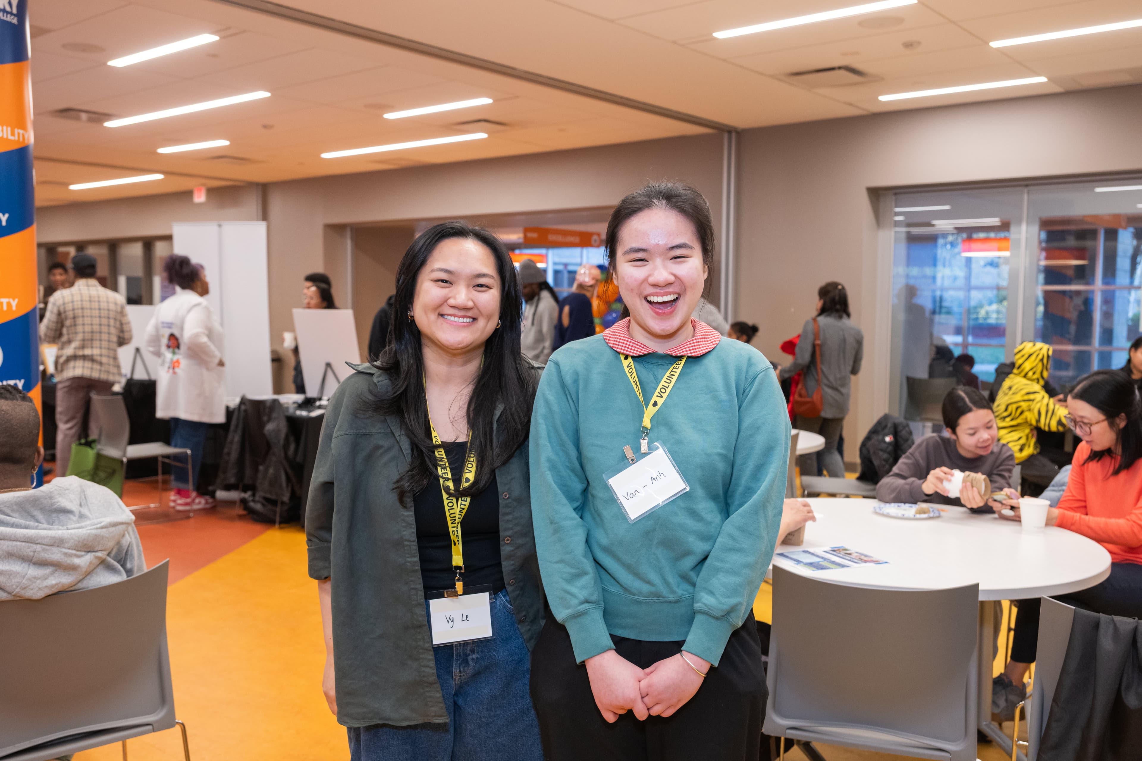 Two young women standing together smiling, both wearing volunteer lanyards, tables and attendees behind them.