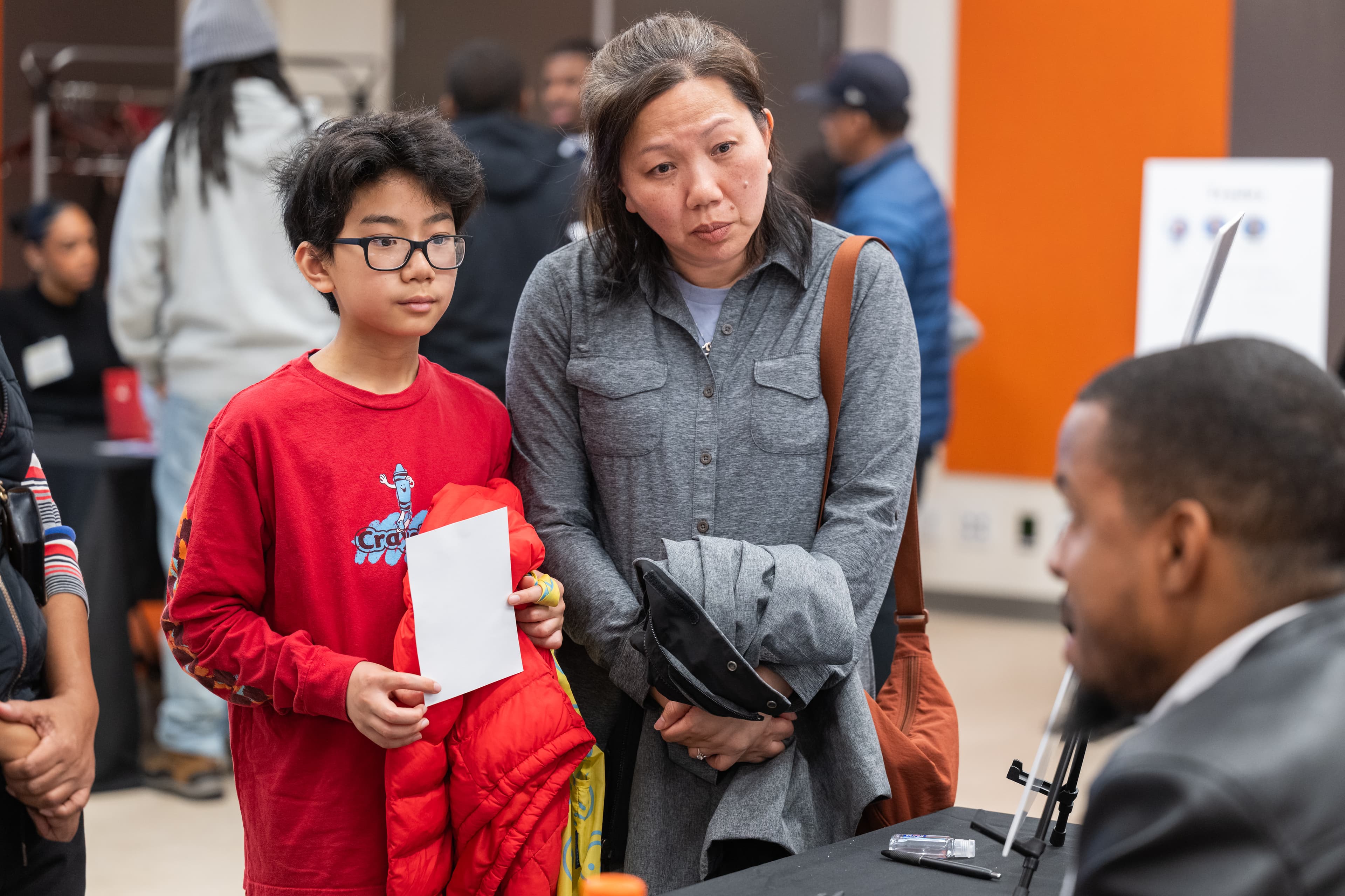 Boy holding a blank card, standing next to a woman looking off-camera, other attendees behind them.