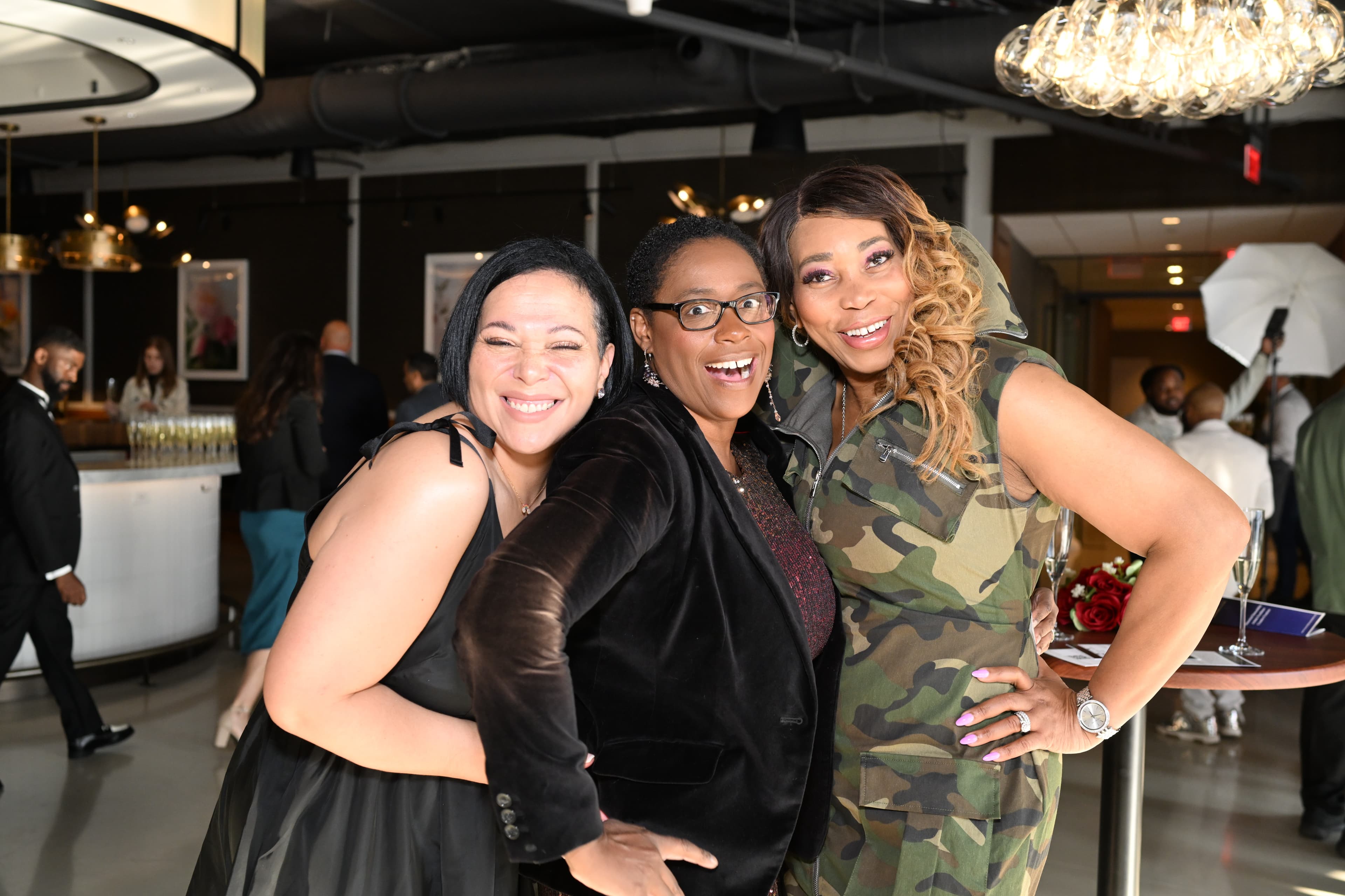 Three women playing it up for the camera, one leaning back with a surprised expression, in a lounge with pendant lighting.