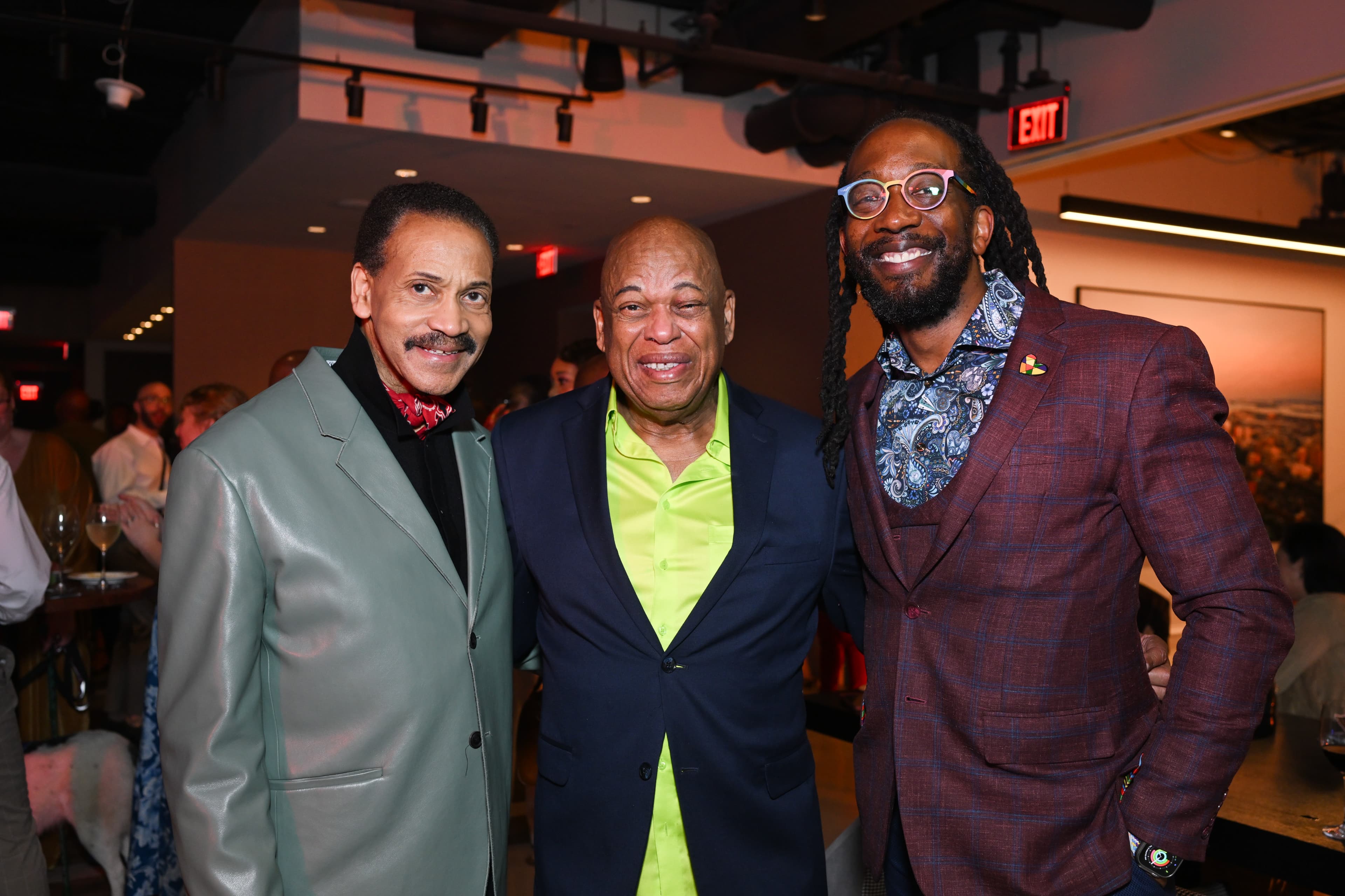 Three men grinning, one in a mint green blazer, one in a bright green shirt, one in a burgundy plaid suit with a rainbow pin.
