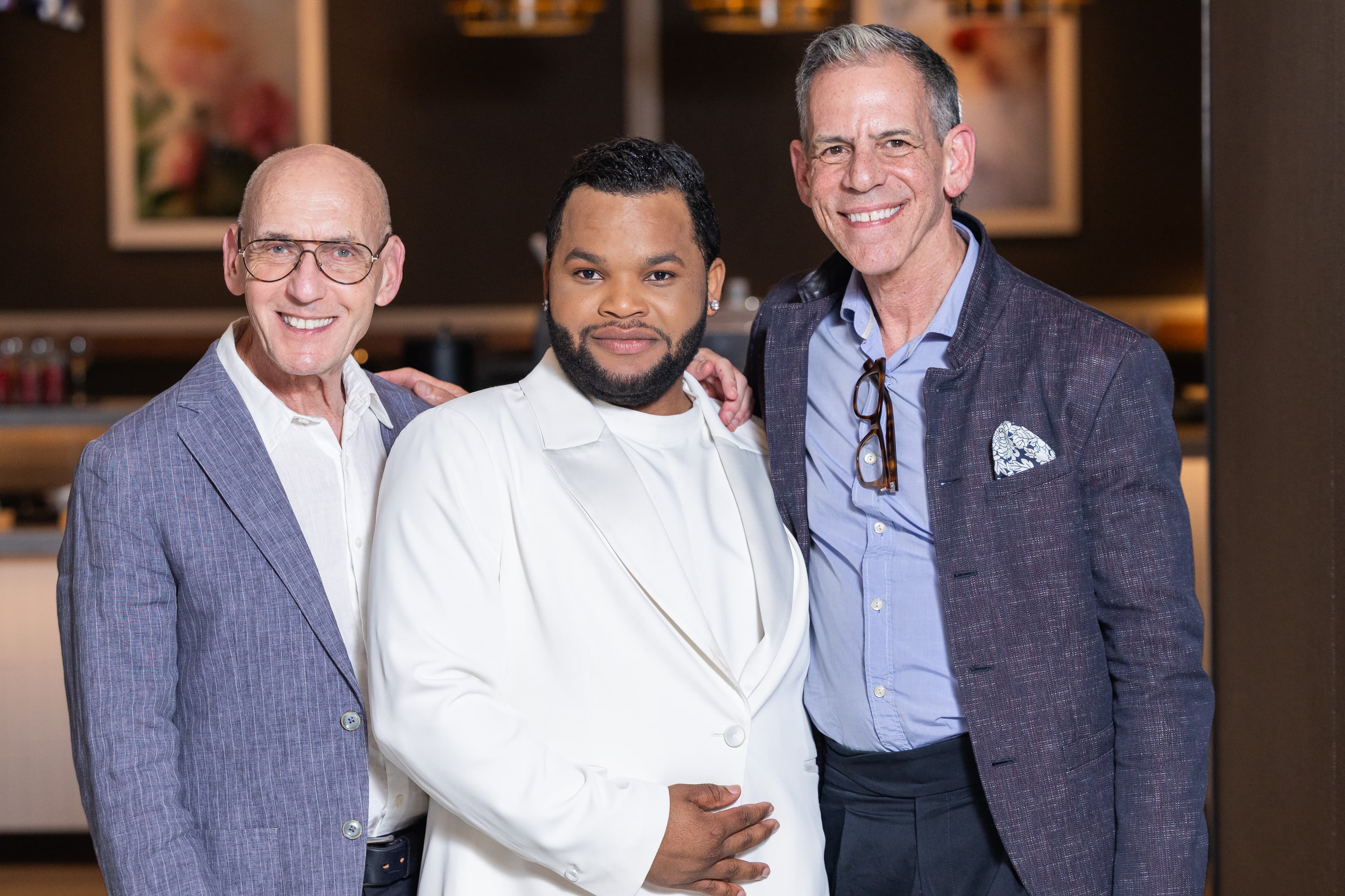 Three men with arms around each other, smiling, one in a white suit.