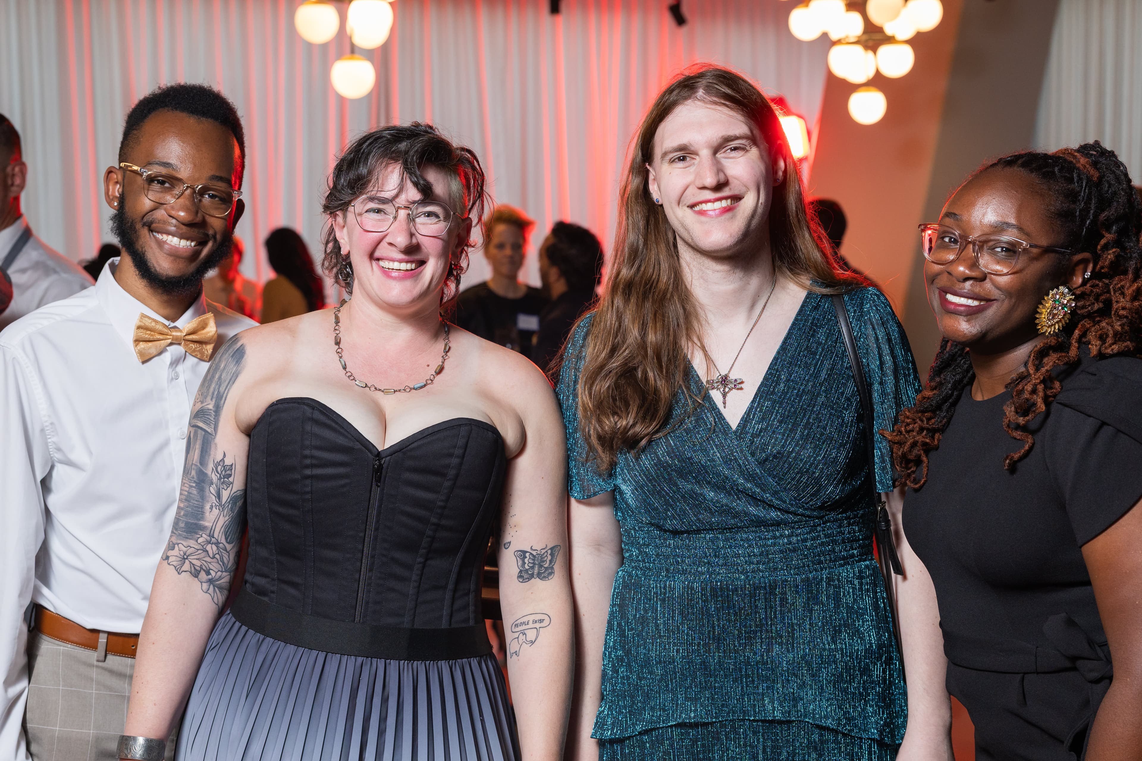 Four people side by side smiling, one with a tattoo on an arm, colorful string lights behind them.