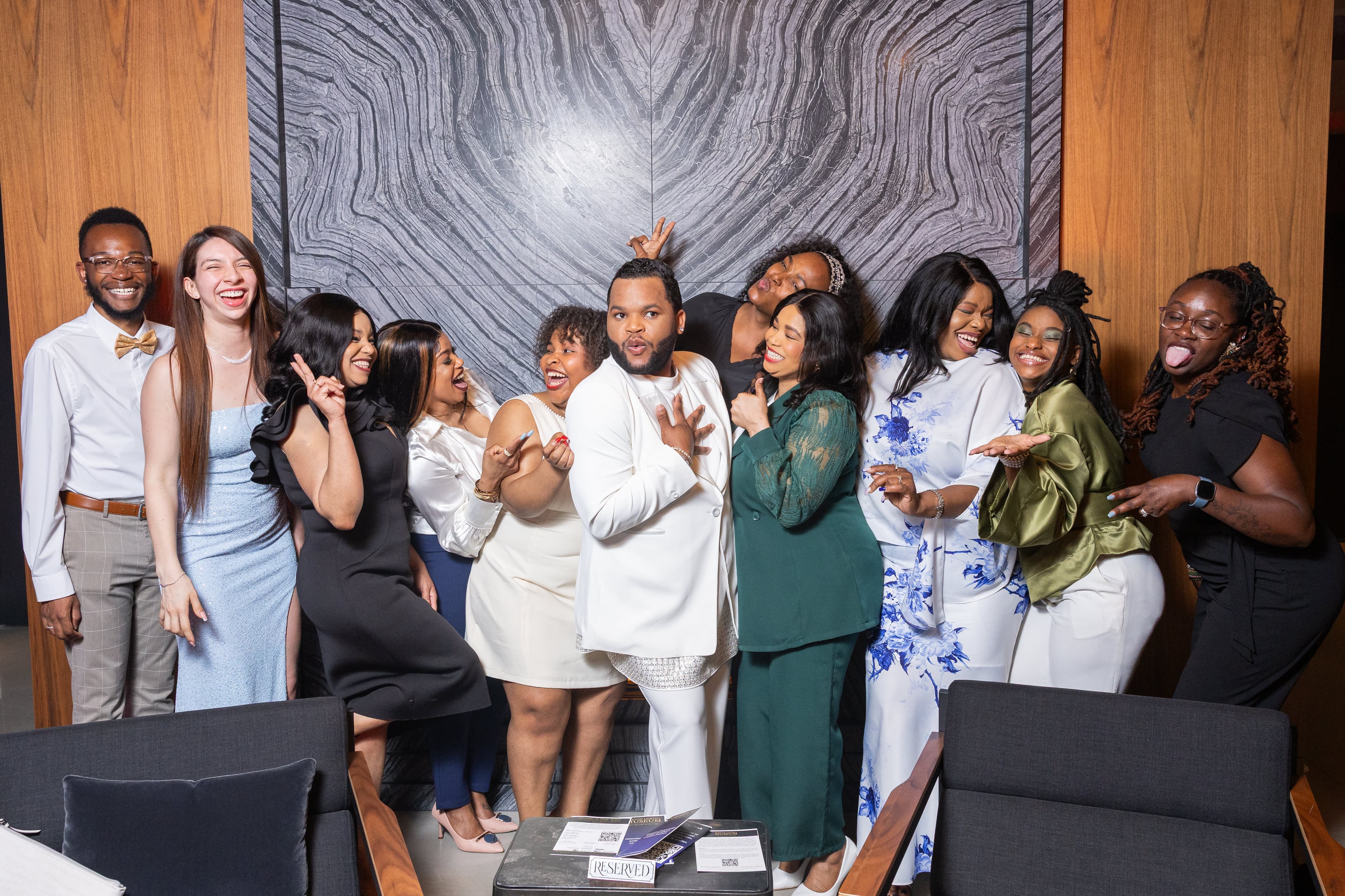Large group of about twelve people laughing and making peace signs together, in front of a marble accent wall.