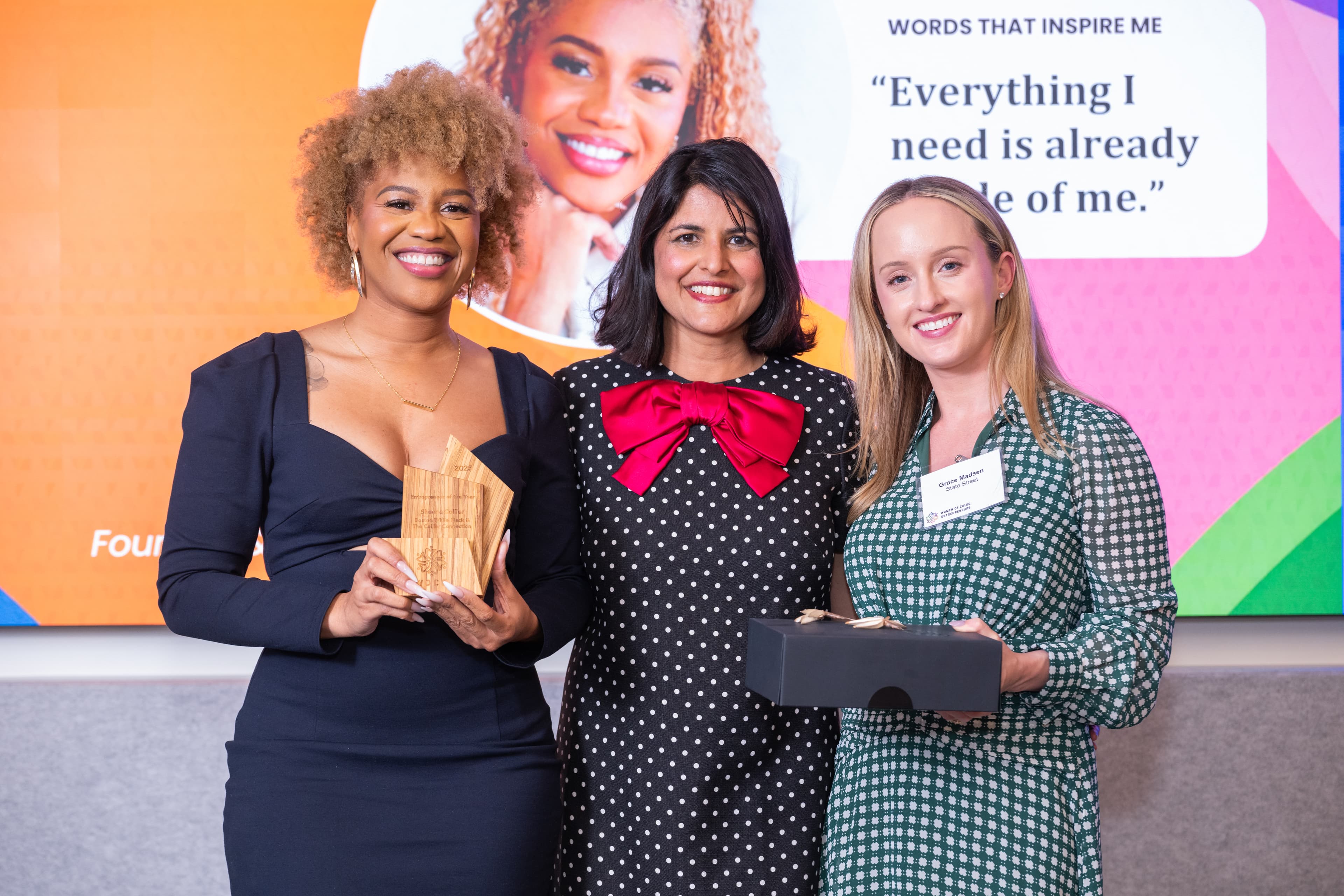Three women standing together, one holding a wooden award, a colorful backdrop behind them.