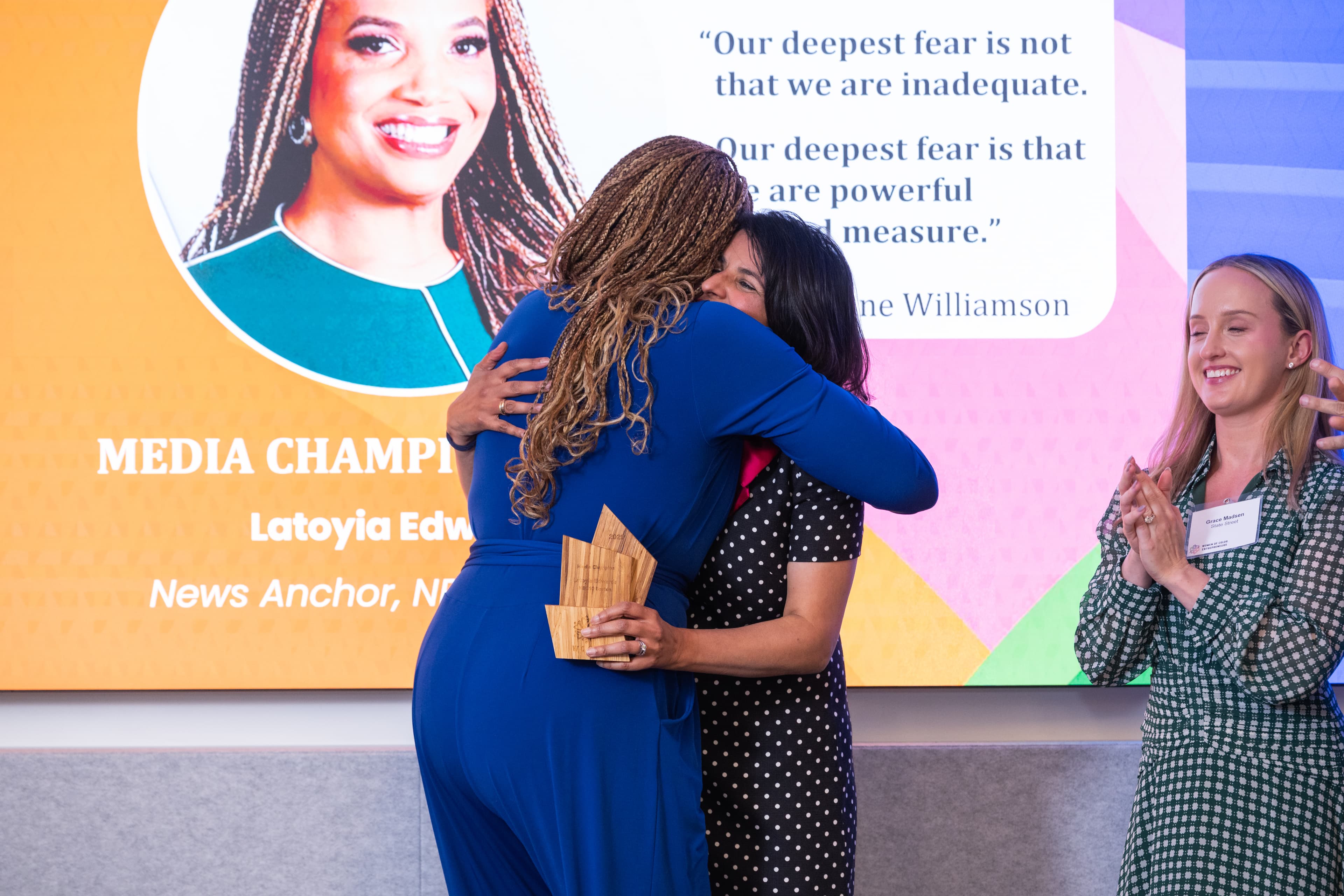 Two women embracing on stage, one holding wooden awards, a third woman applauding nearby.