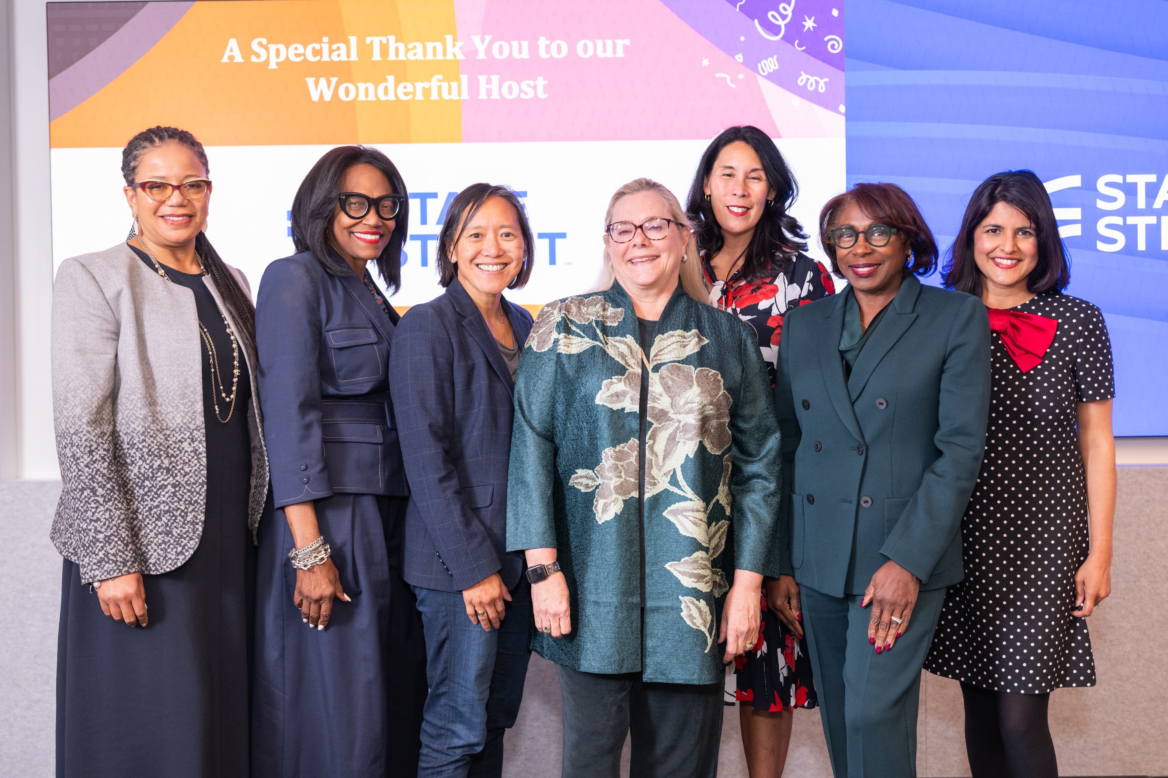 Six women grouped together on a stage, smiling warmly for the camera, a colorful screen glowing behind them.