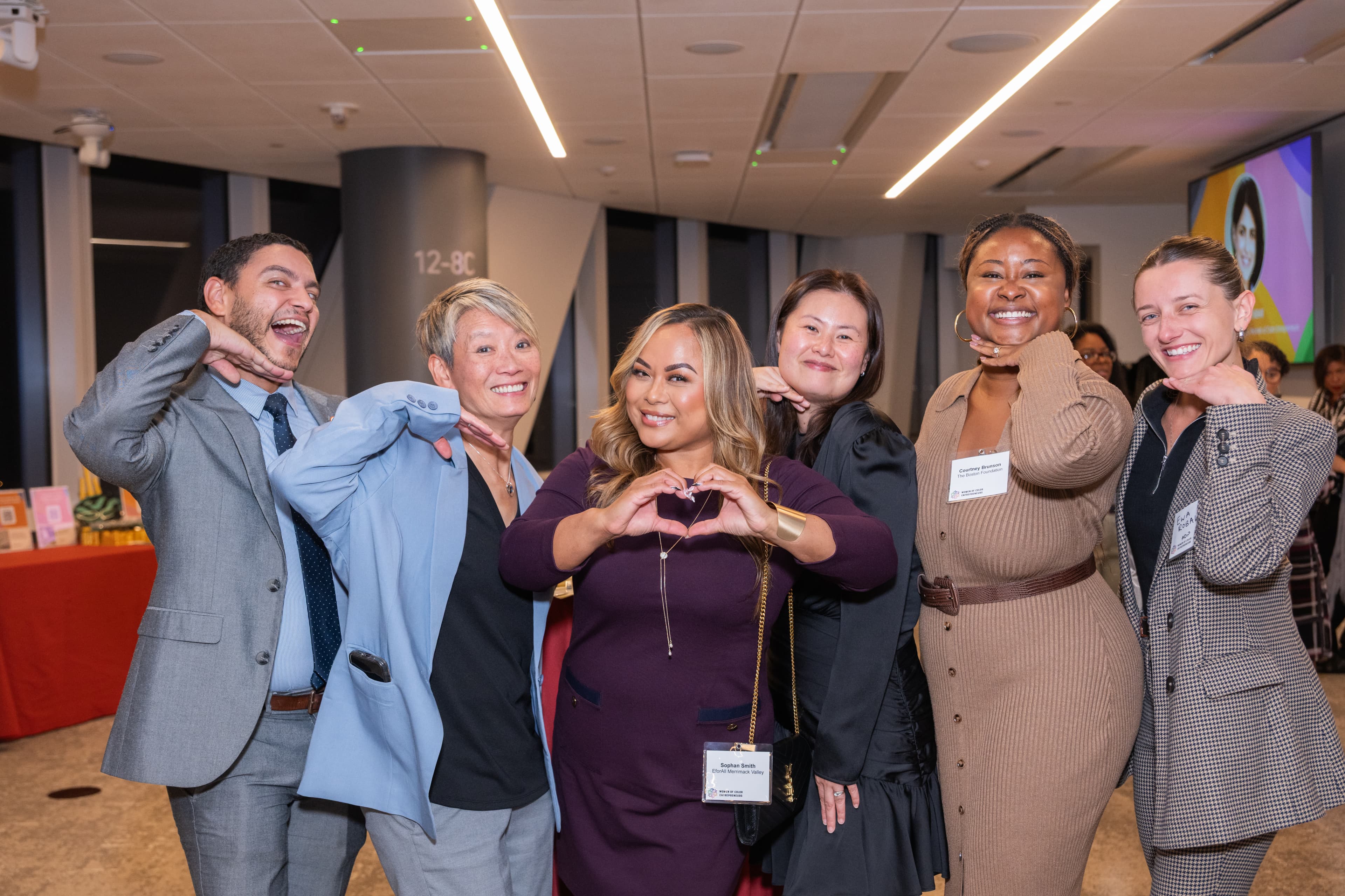 Six people playing it up for the camera, hands near their chins, one making a heart shape with her hands.