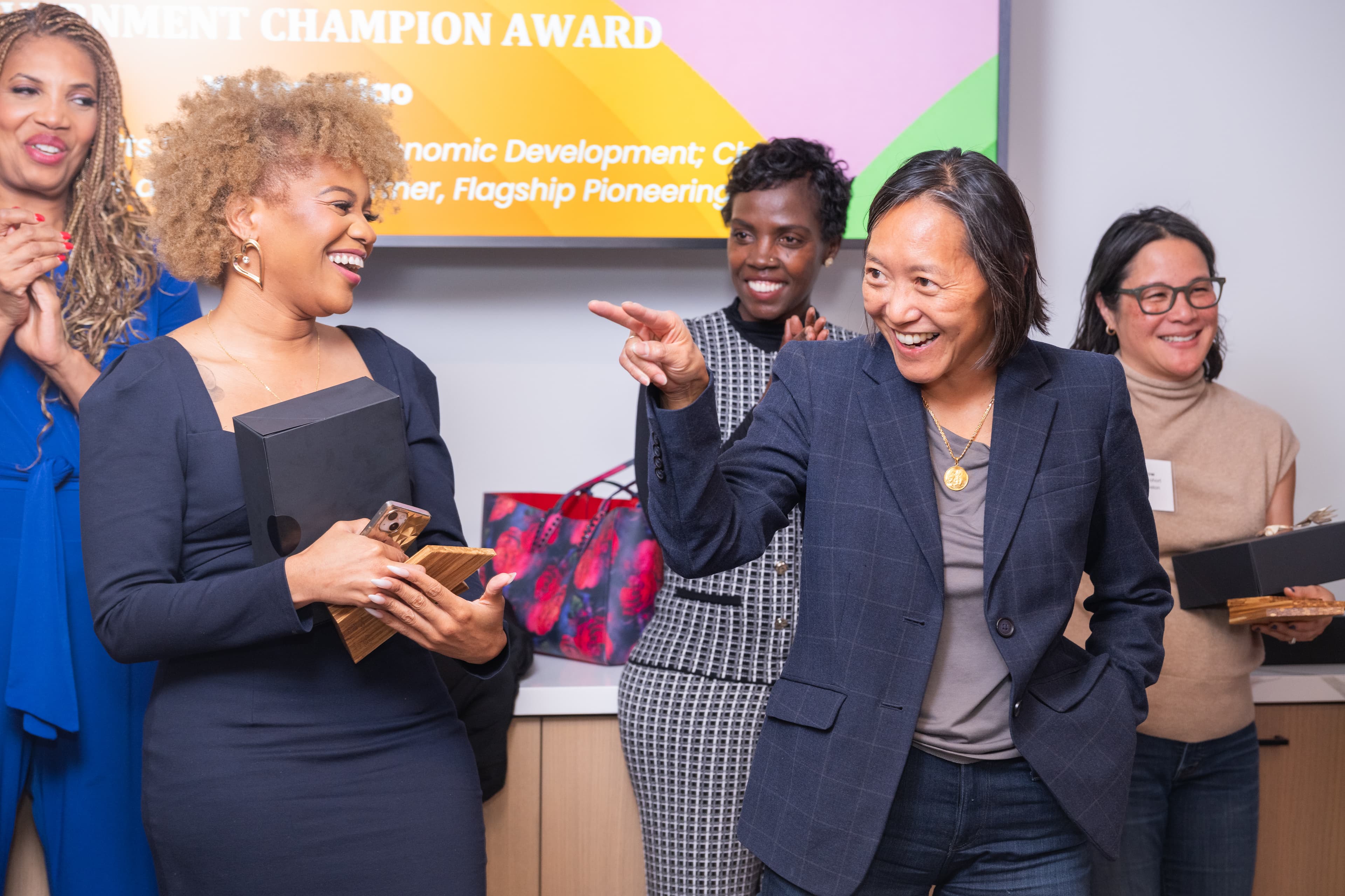 Four women smiling together in a hallway, arms around each other, event signage visible nearby.