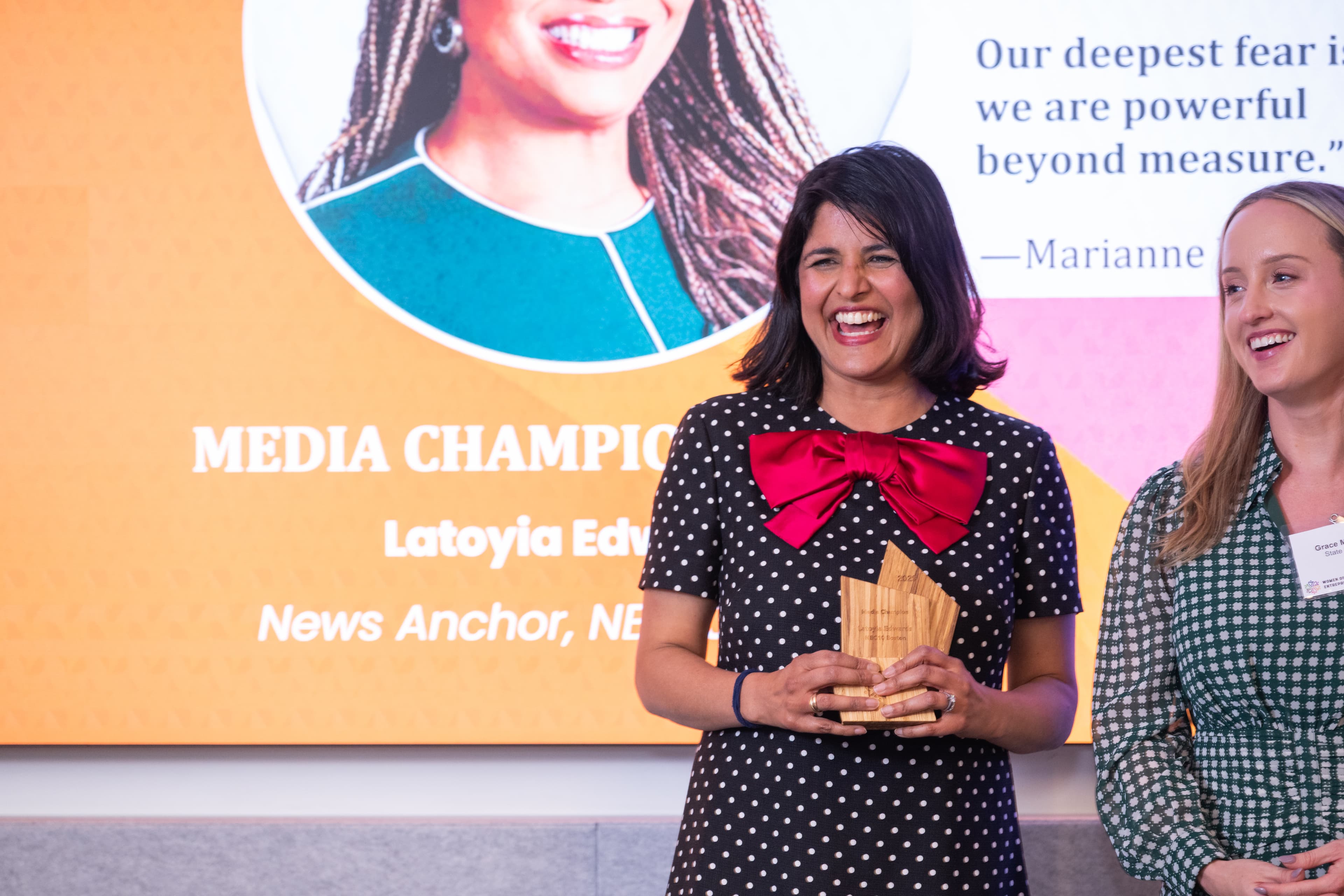 Two women talking and smiling, one holding a wooden award, a colorful display behind them.
