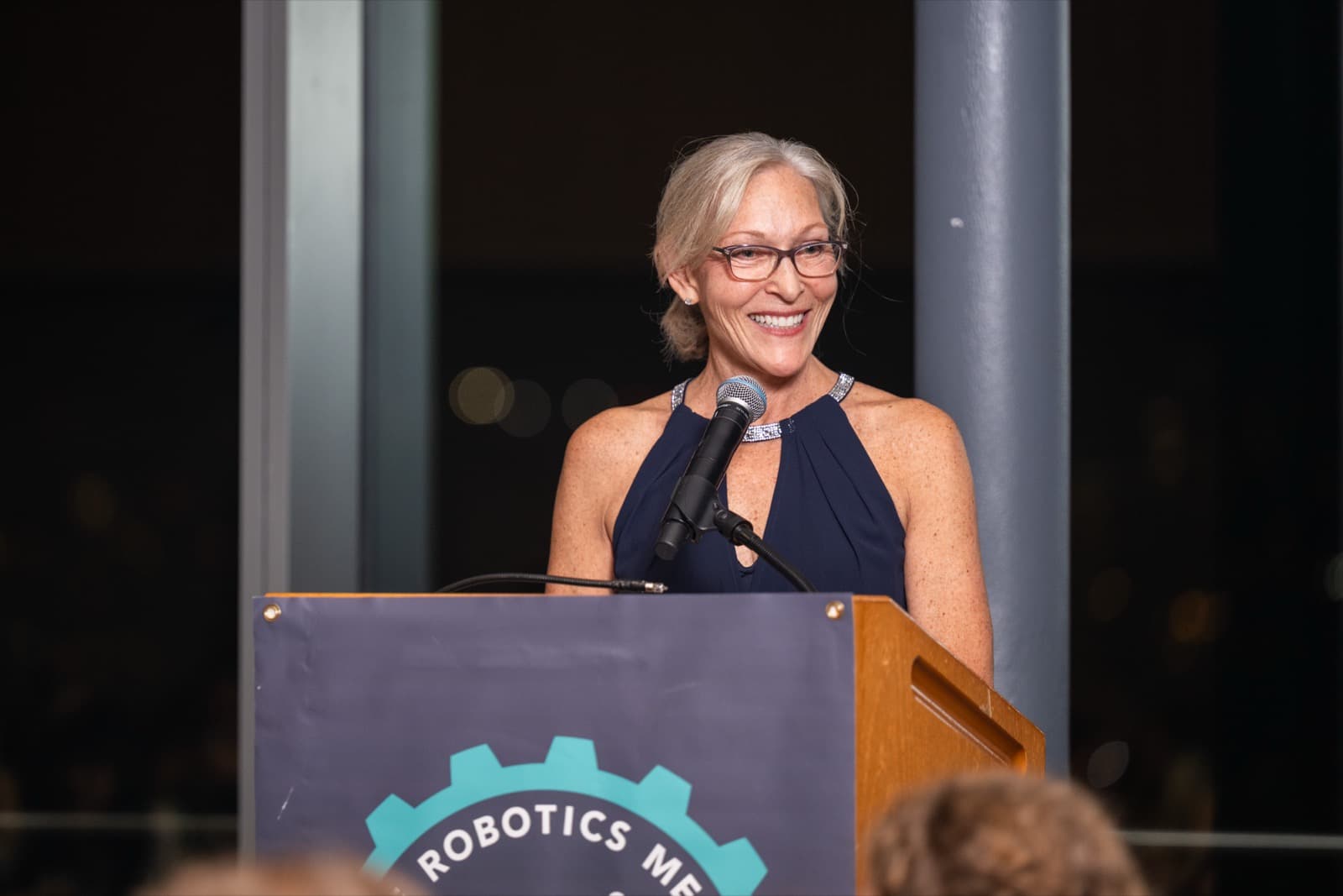 Woman speaking into a microphone at a podium with a teal medal logo, a dark venue with columns behind her.