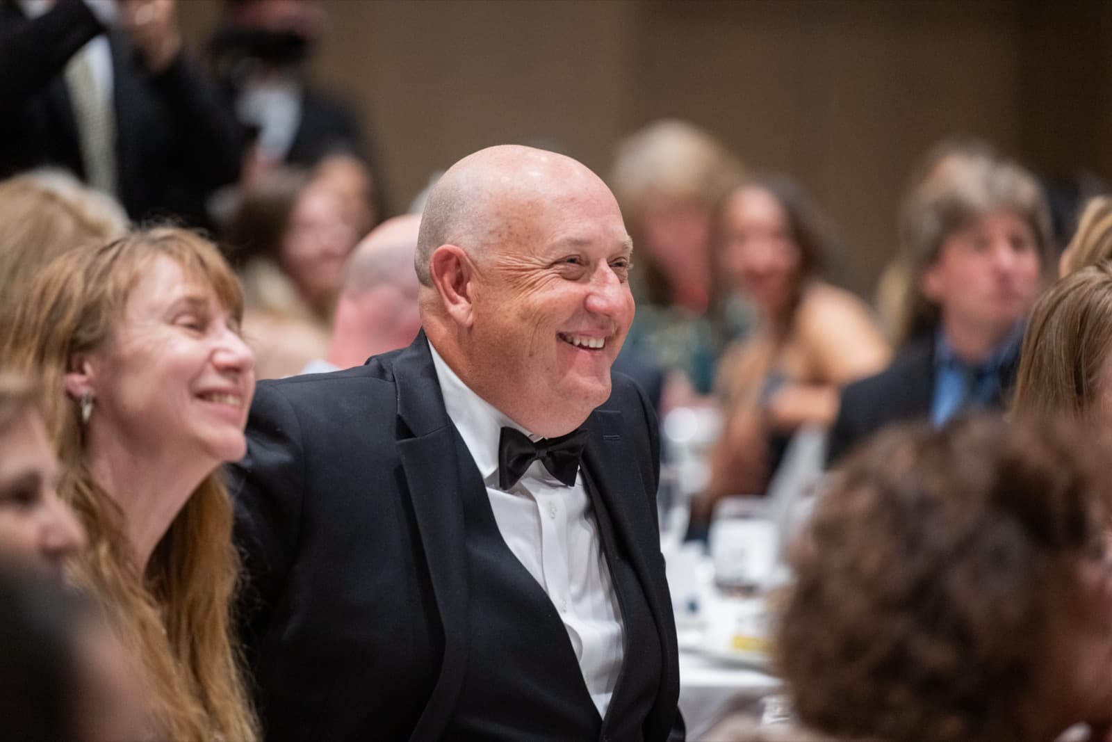Bald man in a tuxedo laughing with a woman beside him at a gala dinner table, other guests blurred behind.