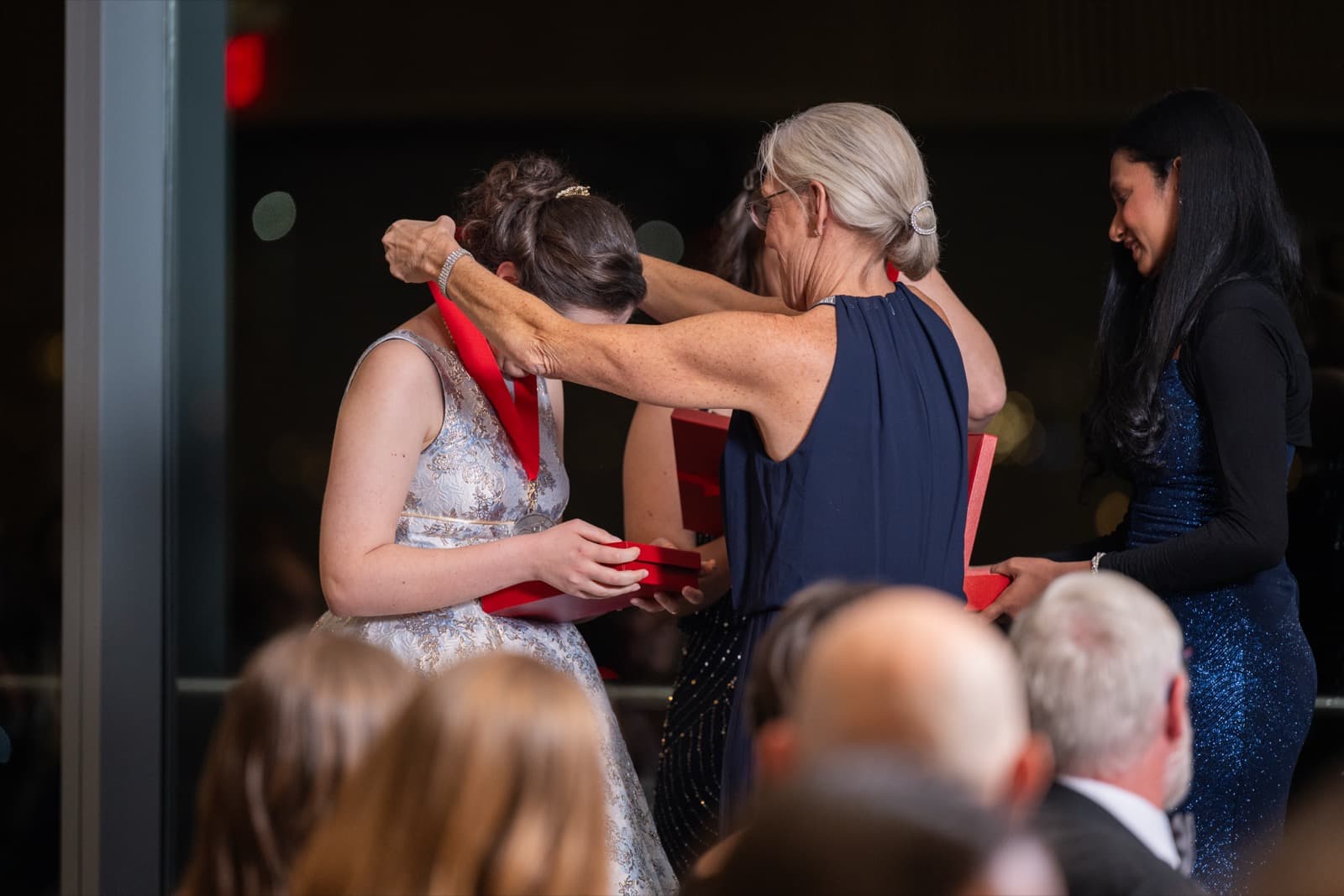 Woman placing a medal on a red ribbon over another woman's head, both in formal gowns, a third woman nearby.