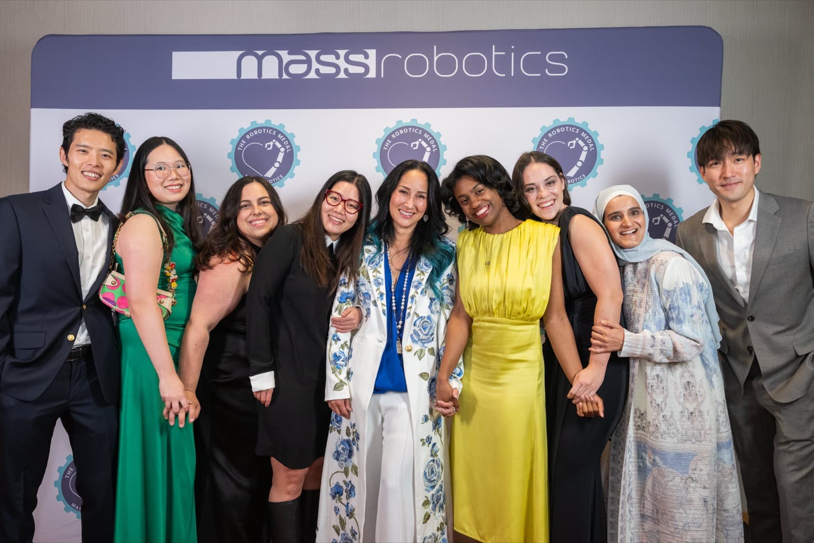 Nine people arm in arm at an event backdrop, dressed in colorful formal wear, big smiles all around.