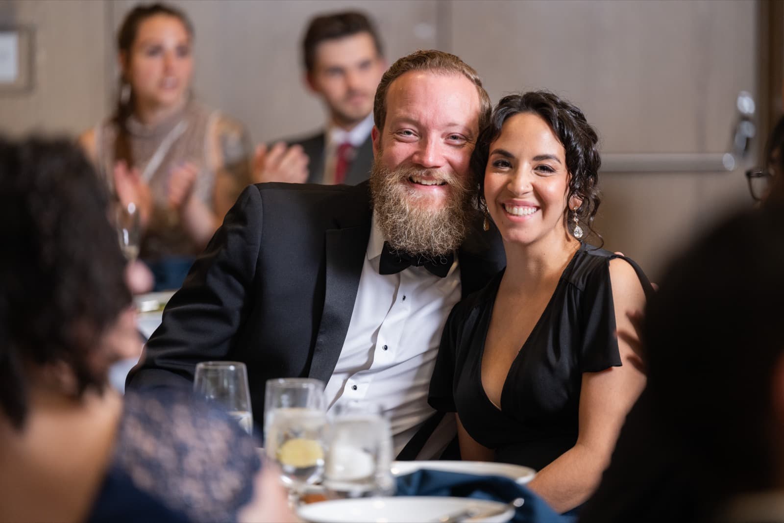 Man with a red beard in a tuxedo smiling cheek to cheek with a woman in a black dress, applauding guests blurred behind.