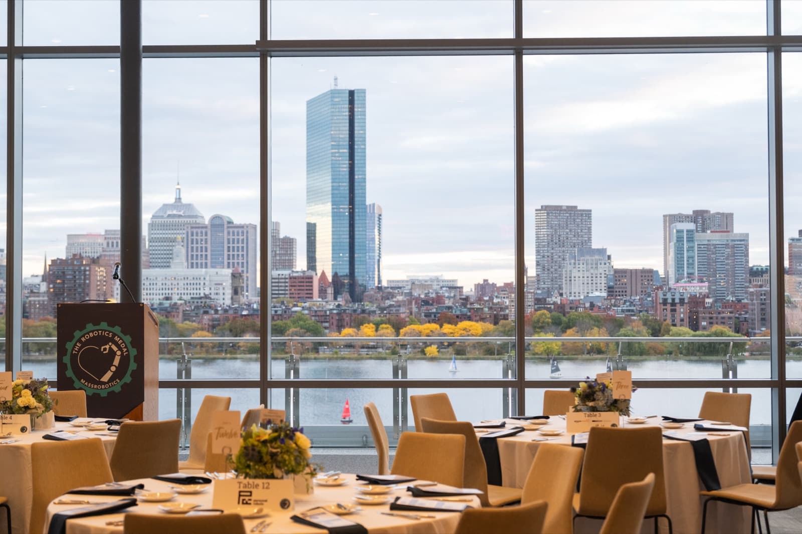 Empty ballroom set for a formal dinner, round tables with floral centerpieces, a podium, a skyline visible through the windows.