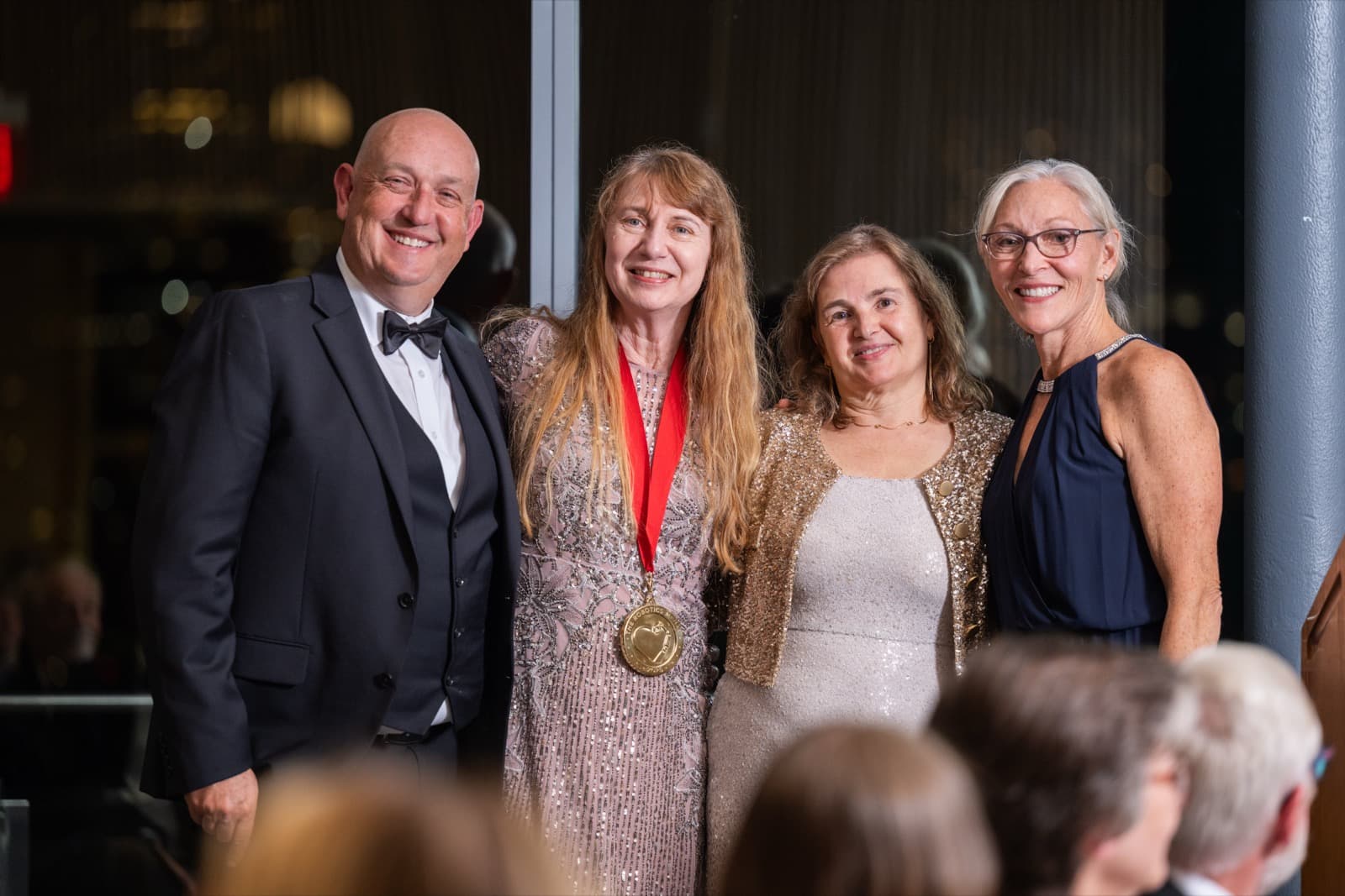 Four people smiling together, one wearing a medal on a red ribbon over a pink sequin dress.