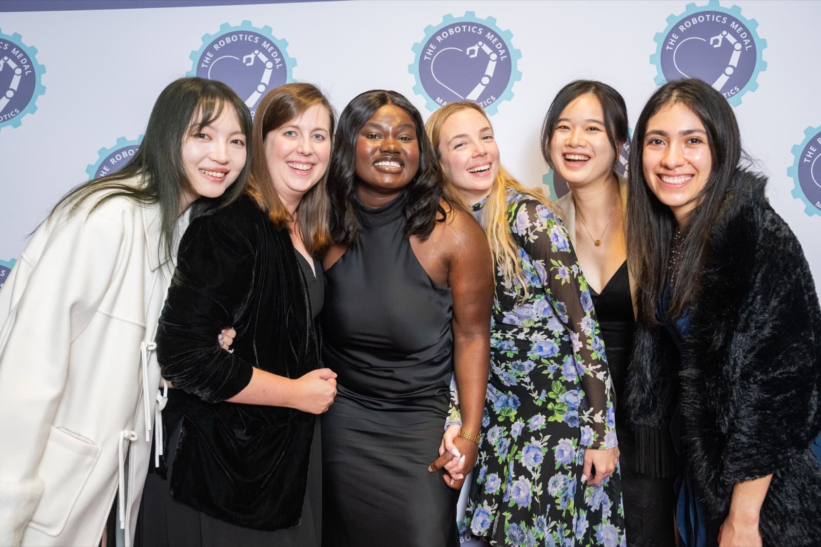 Six women smiling together at an event backdrop, dressed in a mix of black and floral formal wear.