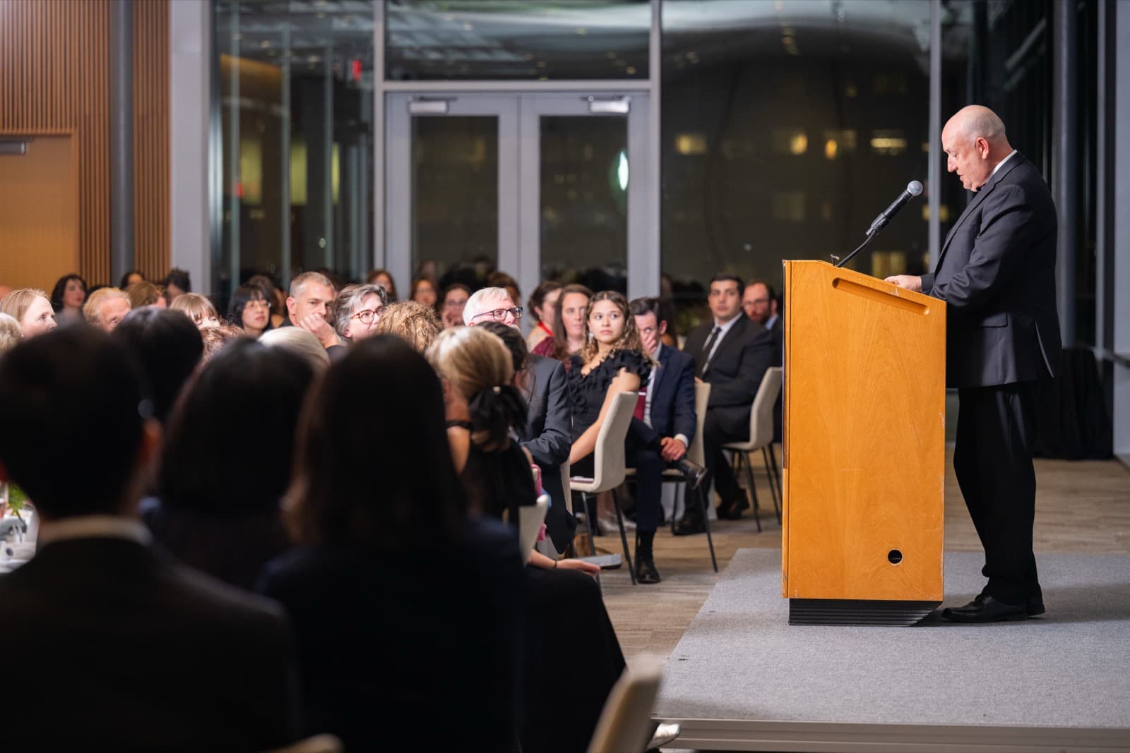 Man speaking at a wooden podium with a microphone, a seated audience listening in a dusk-lit room.