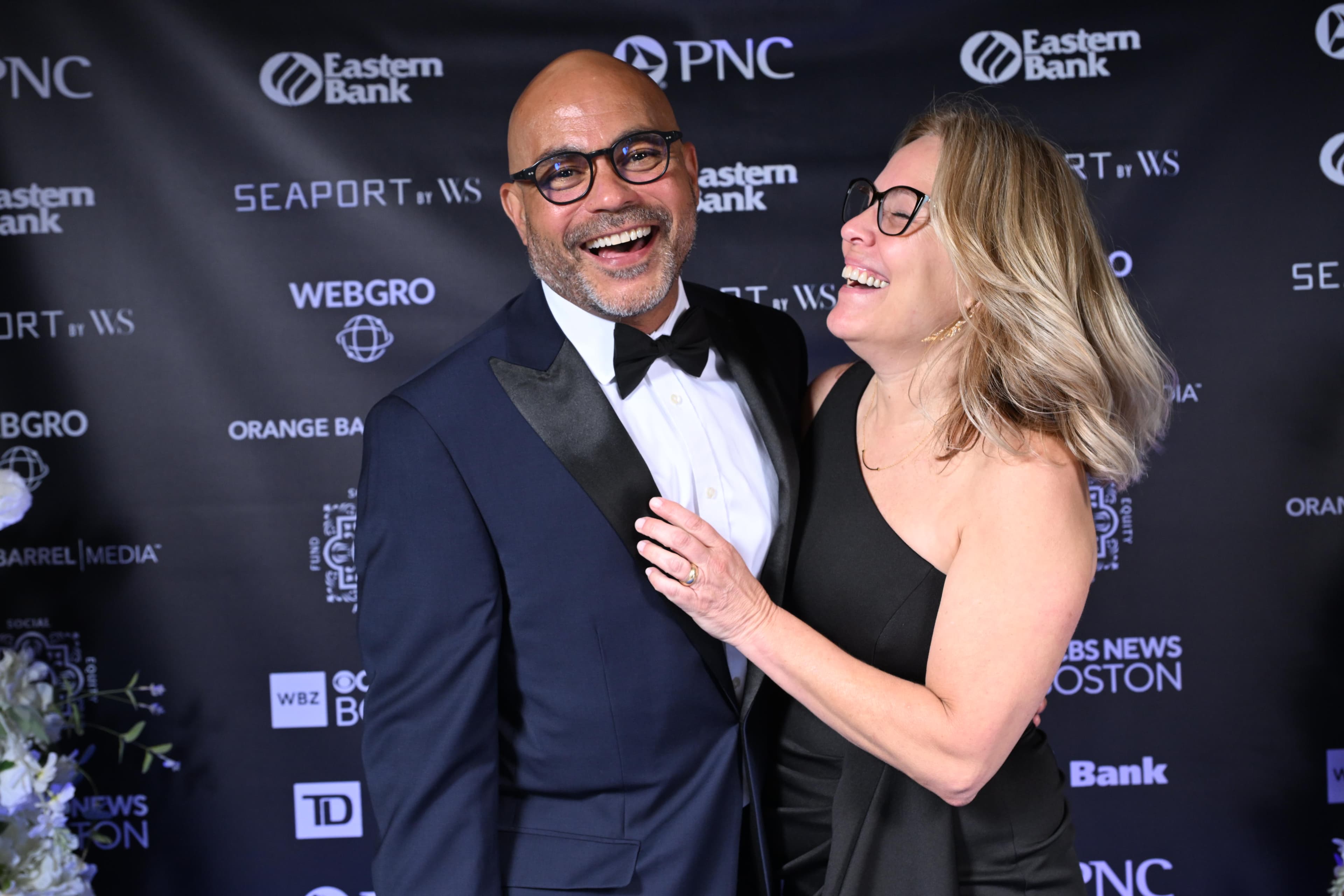 Man and woman laughing together on a red carpet in front of a sponsor step-and-repeat.