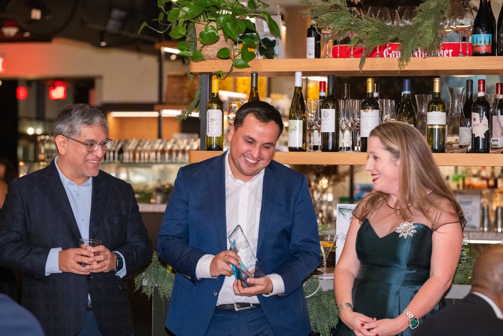 Three people smiling together, one holding a glass award, wine bottles on shelves behind them.