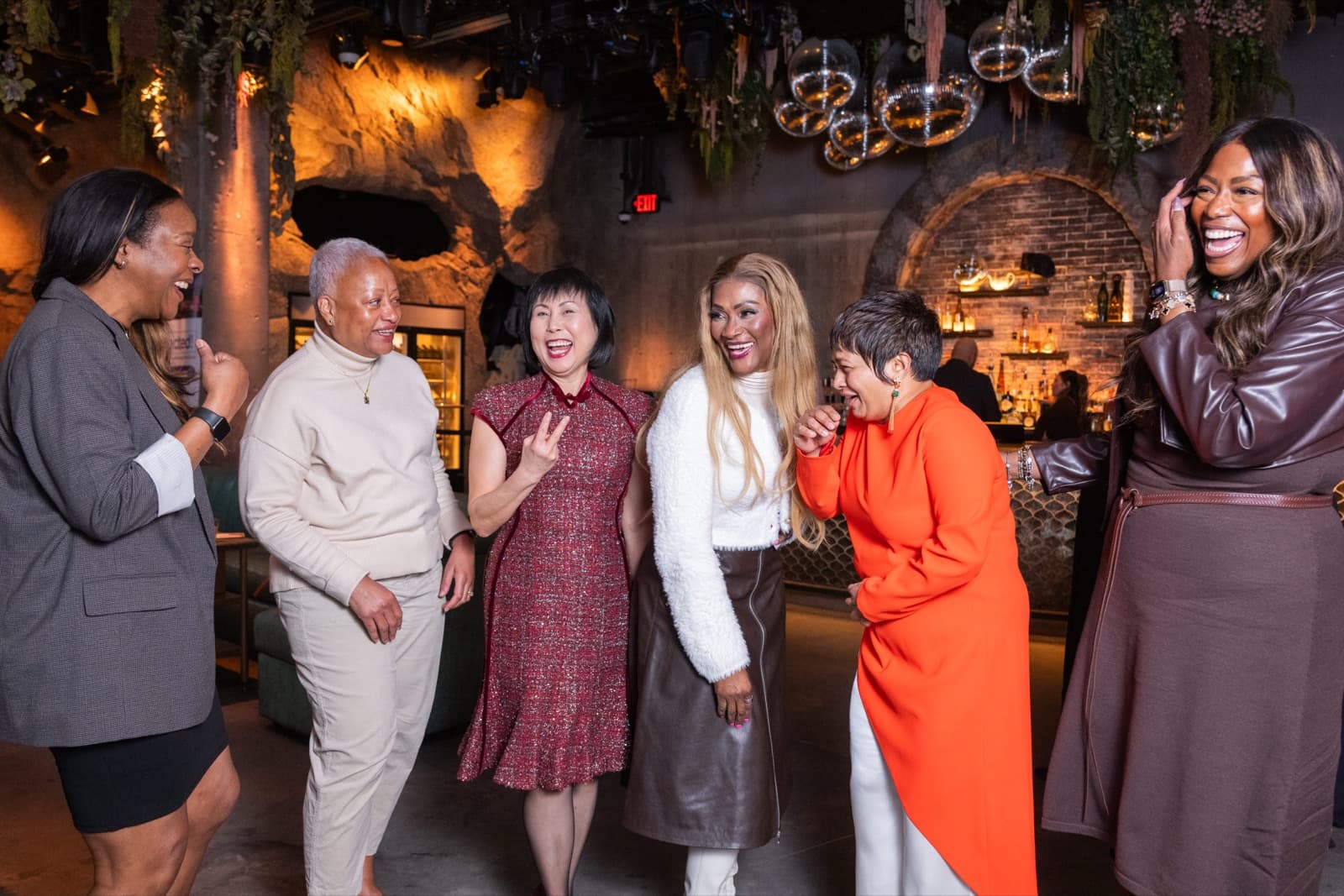 Six women laughing together at an indoor lounge, disco balls and greenery hanging above them.