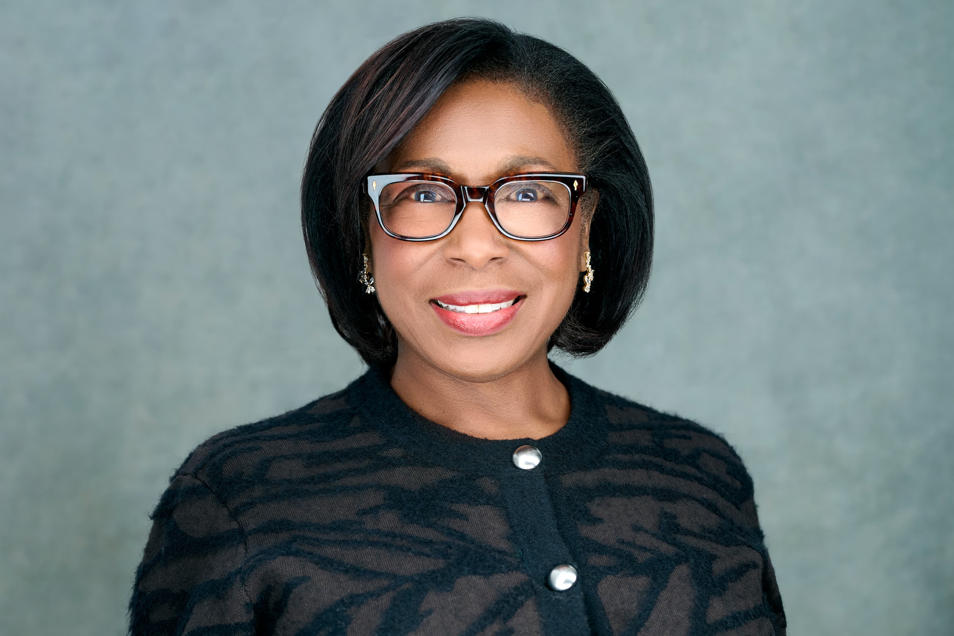 Woman with a black bob and tortoiseshell glasses smiling, sage-green studio background.