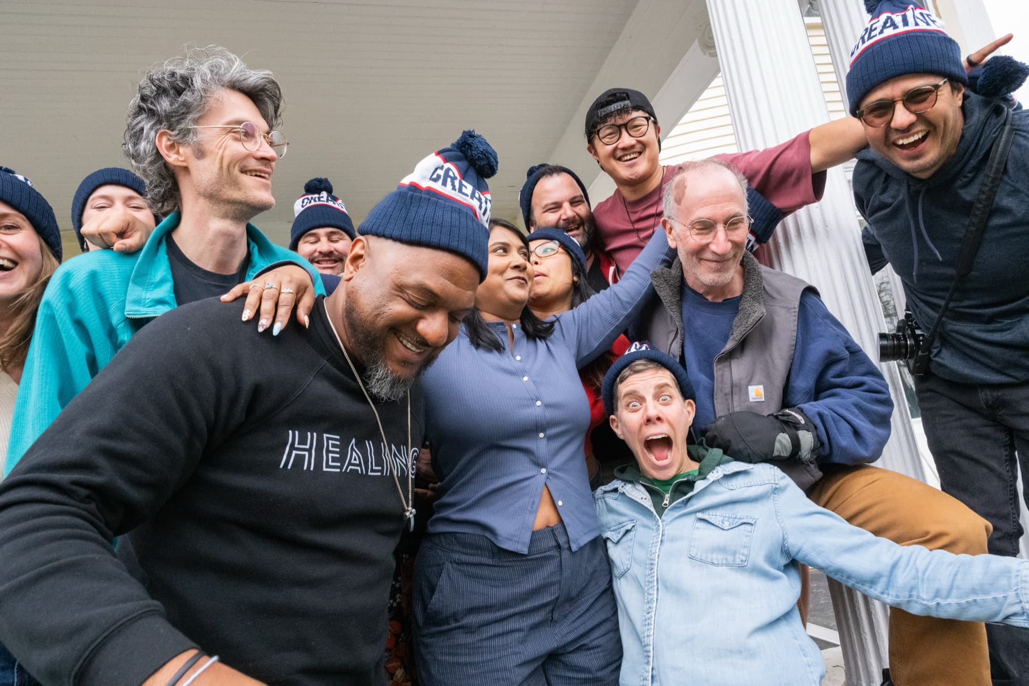 Large group posing playfully together on a porch, many wearing matching knit beanies, laughing.